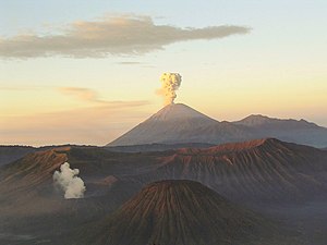 A brown volcano in the centre with white smoke emanating from its peak, a cloudy sky fading from blue at the top through yellow in the middle to red at the horizon, and brown mountains in the foreground.